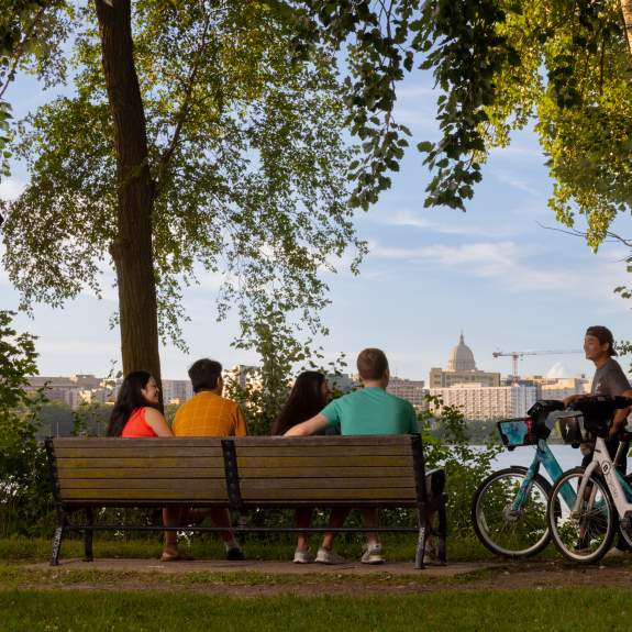 A shot of the city skyline over the lake while a group of people sit on a bench and two others stand next to them with bikes overlooking the views.