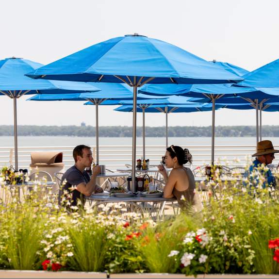 People sitting at an outdoor restaurant that over looks the lake and is surrounding by flowers and greenery.