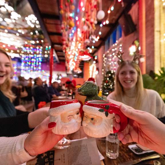 A group of women at a restaurant decked out in Christmas lights, cheersing their Santa mugs