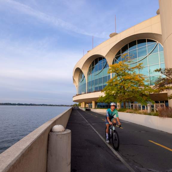A man in a biker suit wearing a helmet as he rides his bike past the lake and Monona Terrace Convention Center.