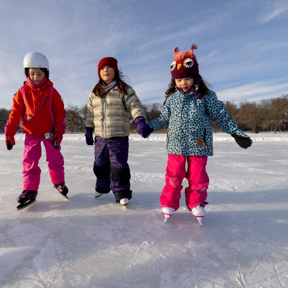 Three young girls are bundled up for winter and are holding hands while ice skating on the frozen lake.