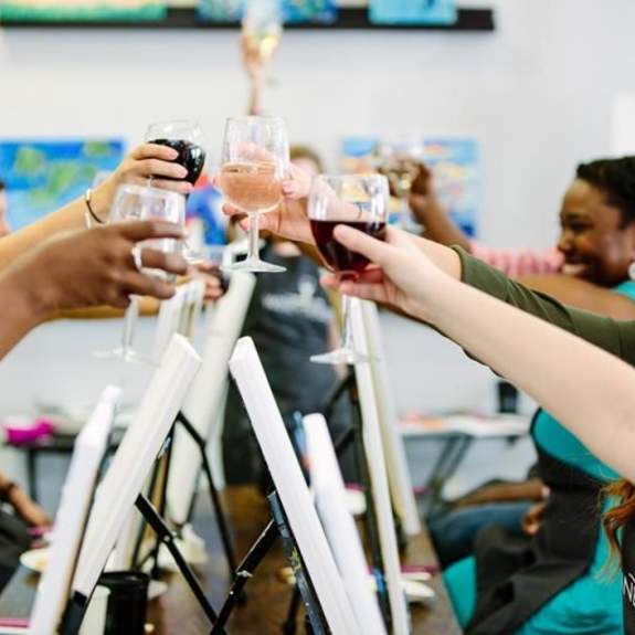 A group of women clinking wine glasses over canvases at a wine and paint event