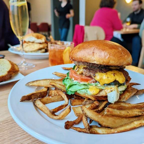 A large burger with cheese, tomato, lettuce and sauce served on a white plate with fries. An old fashioned and glass of champagne are also on the table.