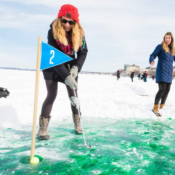 A woman puts a bright yellow golf ball into an indentation on a frozen lake. The indentation is marked with a flag indicating a mini putt course on the frozen lake. Two bystanders cheer her on