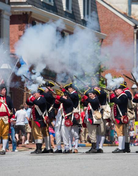 A group of historical reenactors in colonial military uniforms fires muskets, creating clouds of smoke during a lively outdoor event.