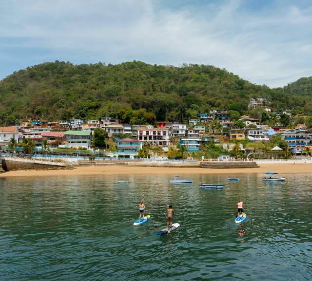 Three people practice paddleboarding in calm waters near the beach and colorful houses on the hillside of Taboga Island, surrounded by lush vegetation.