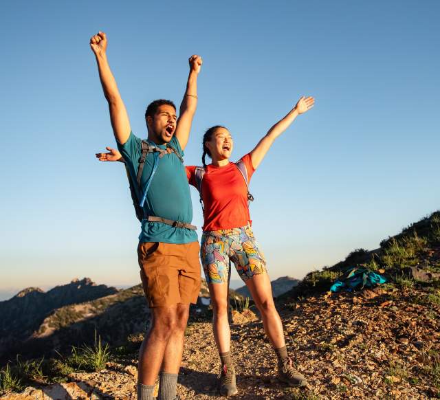 Man and Woman wearing hiking gear with their arms up and looking excited to reach the top of a mountain