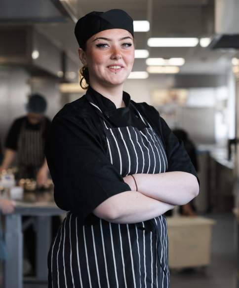 A chef standing with their arms folded and smiling in the centre of a commercial kitchen.