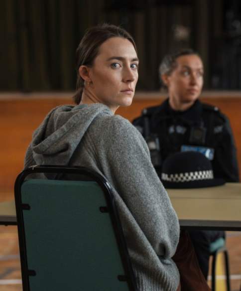 A woman in a grey hoodie sits at a table in a large hall, looking towards the camera. Two uniformed police officers sit across the table, with two police hats placed in front of them.