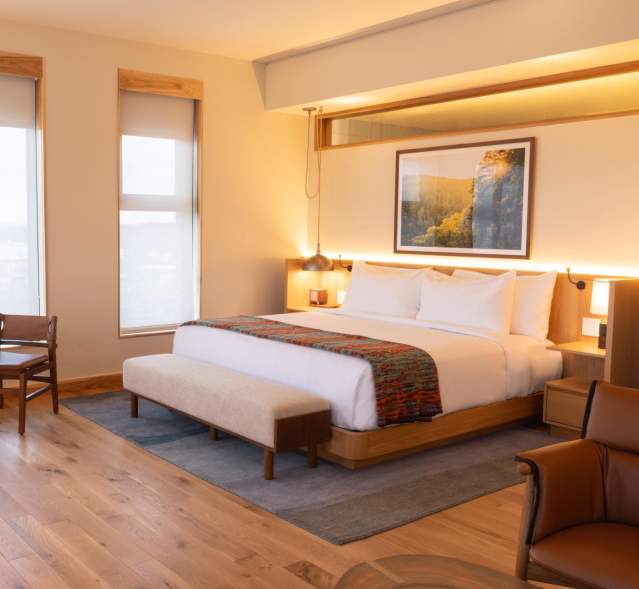 Modern hotel room featuring a king-sized bed, wooden furniture, and large windows with natural light. A small dining table and chairs are present.