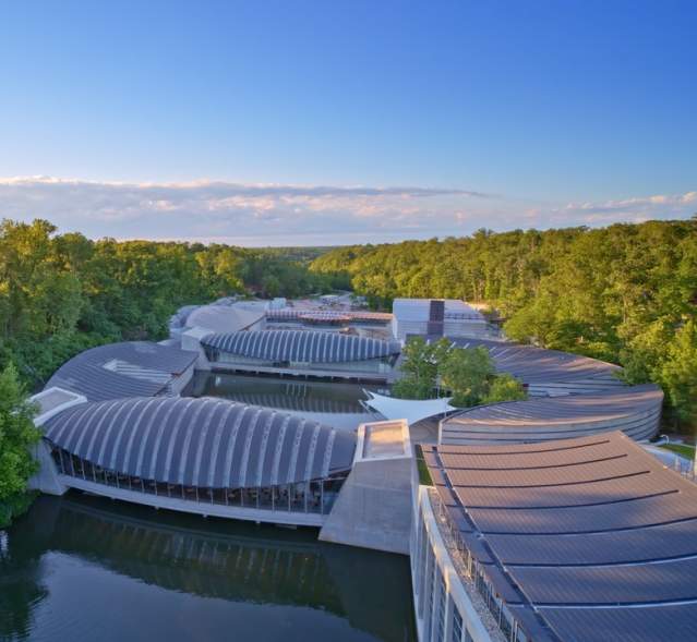 Aerial view of modern architectural structures nestled among lush greenery, reflecting on a serene waterway under a clear blue sky.