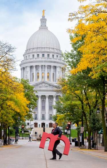 A man carries a giant "M" across a street with the Capitol building in the background
