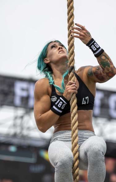 A muscular white woman with bright blue hair climbs a rope outdoors at the CrossFit games.