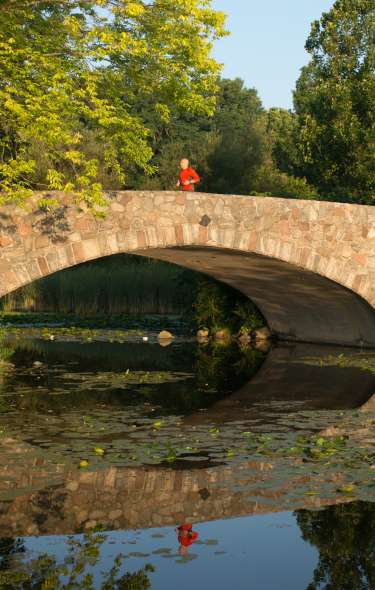 A wide view of a person running on a stone bridge that crosses over a pond.