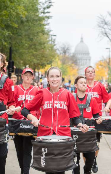 Members of the University of Wisconsin marching band, led by the drum line, march on downtown street with the Capitol building in the background.