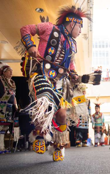 A man wearing traditional Native American clothing does a dance on stage in front of a crowd of dozens of people