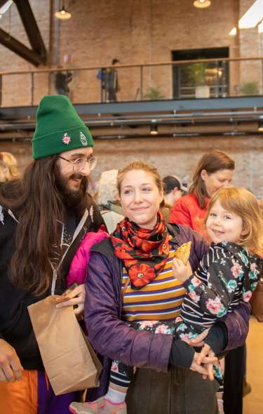 A man, woman, and small child shop the indoor Late Winter Farmers Market at Garver Feed Mill. They have shopping bags in hand and are surrounded by vendors selling microgreens