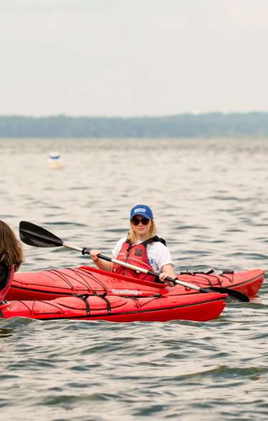 Two women kayak on a lake. Three other boats are sailing in the background