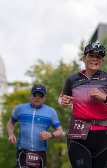 A white woman and a white man during the run course of IRONMAN Wisconsin. Running down State Street with the State Capitol behind them.