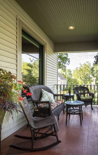 A front porch overseeing a wooded area. On the porch are hanging flower baskets and wicker furniture