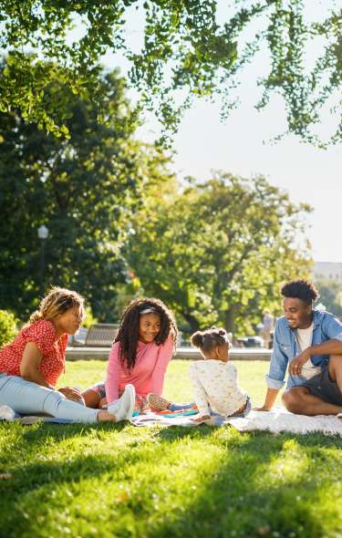 A Black family of four people enjoy a picnic on the lawn in front of the Capitol building on a sunny day. The two adults are smiling at the two children.