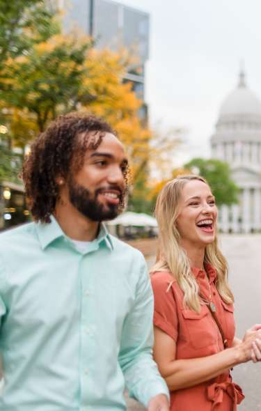 A Black mand and a white woman are laughing while looking at something out of frame. They are standing on King Street in Downtown in front of a row of outdoor dining establishments with front facing patios. The Capitol building is in the background.