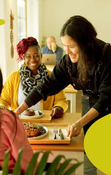 A woman sets a tray of food down on a table between two women