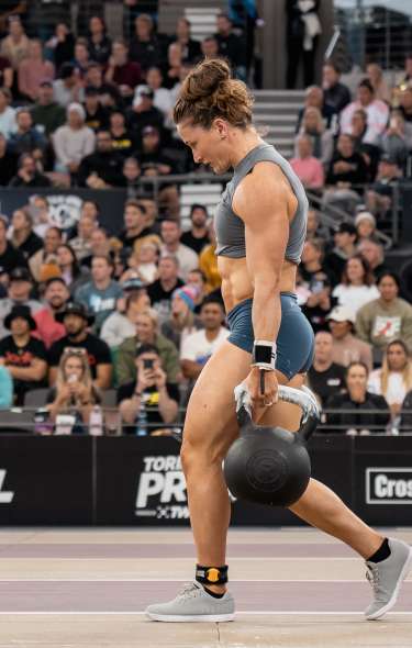 A woman walks with kettlebells in each hand at the CrossFit Games. A crowd of spectators are in the background.
