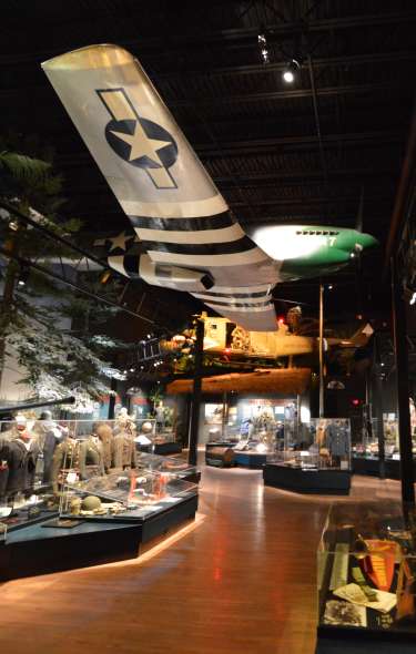 A section of a museum featuring historic military uniforms. A small military airplane hangs from the ceiling above the exhibits