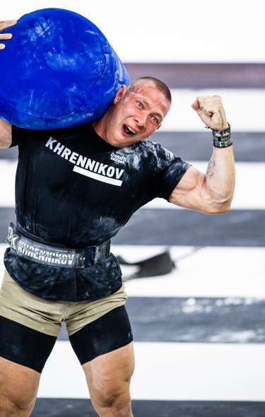 A muscular white man holds a bright blue sandbag on his right shoulder and flexes his left bicep at the CrossFit games. He's smiling triumphantly.