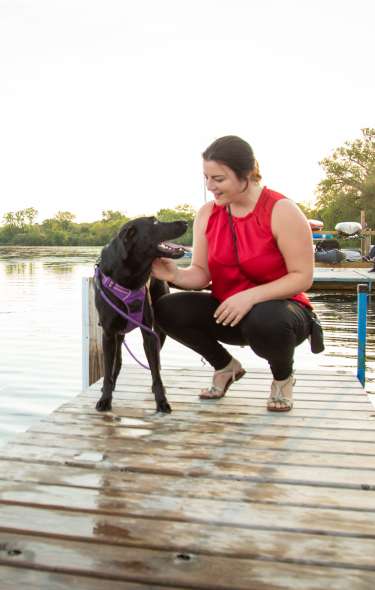 A woman pets her Black Labrador dog on the pier. A group pf boats are docked behind them