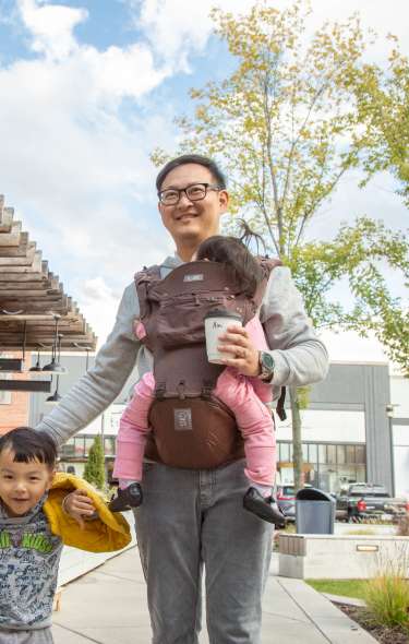 An Asian man walks down the street carrying coffee and holding a young boy's hand next to him. He has a baby in the front carrier strapped to his body