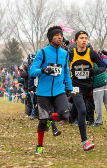 A group of kids cross-country run in cold weather. Two kids are pulling ahead of the crowd and spectators are lining the path.