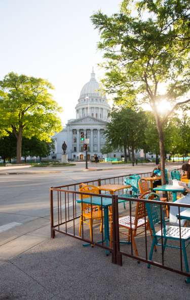 A wide shot of the Wisconsin State Capitol building in the distance, the sun peaking through the trees and people sitting on a colorful patio outside a coffee shop.