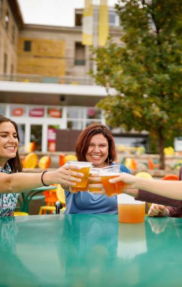 Three young adult women and one adult man cheers with beer glasses while sitting on the sunburst chairs at Memorial Union Terrace. They are smiling and laughing.
