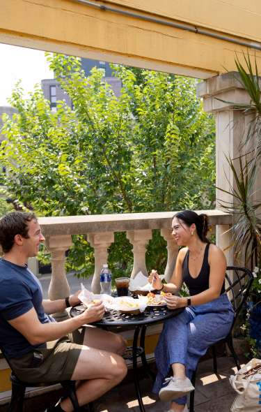 Two tables with two people at each sitting and enjoying their food on the rooftop patio at a local restaurant on a sunny day.