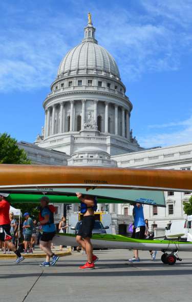 Groups of two people at a time carry canoes over their heads in front of the Capitol building. People are dressed in swim and summer attire