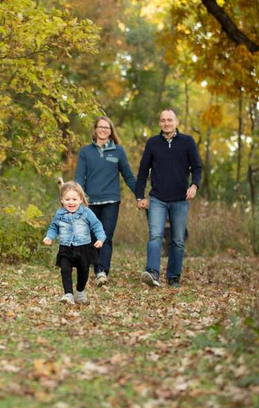 Two adults hold hands while a young child runs ahead of them as they hike through a wooded forest during Fall