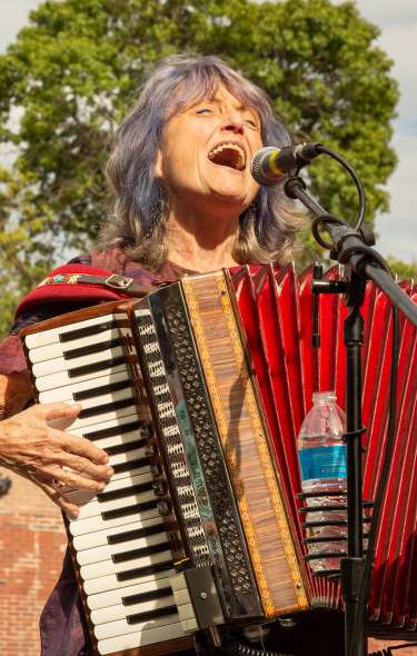 A woman sings while playing the accordion outdoors in front of a building. The sky is full of clouds.