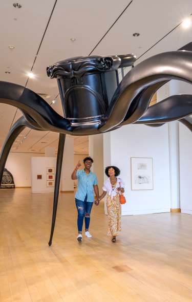 Two people walk through an art gallery under a giant spider-like sculpture