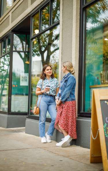 Two women are talking on the sidewalk of Ancora Coffee Shop in a Downtown setting. A menu board in front of them shows off specials of the day.