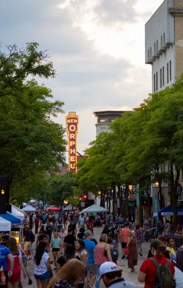 A crowded city street lined with vendor tents and trees, with people walking between historic buildings and the glowing New Orpheum Theater sign in the background at dusk.
