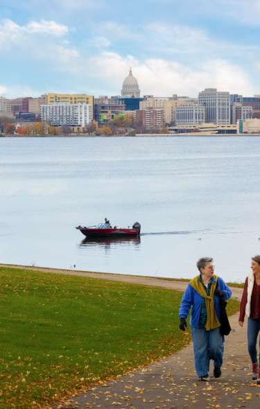 Two people walk along a lakeside path covered with fallen leaves, with Lake Monona and the Madison skyline in the background.