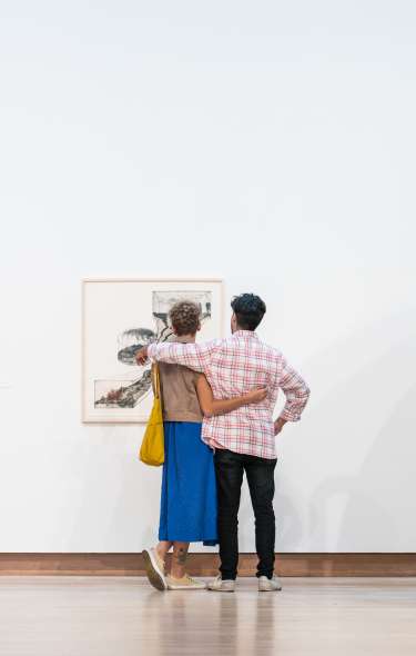 Two people look at a a series of paintings on a wall as they wrap their arms around each other.