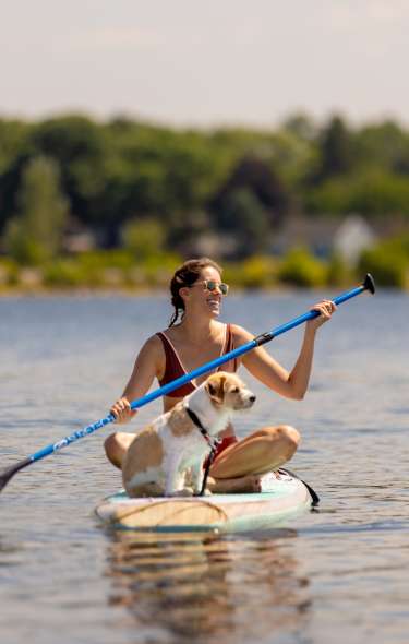 A white woman and a medium sized white and brown dog sitting on a paddle board in the lake with rows of trees behind them.