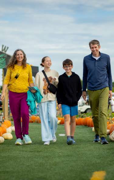 A group of four people walks along a path lined with pumpkins and gourds at a pumpkin patch. A decorative farm entrance sign is visible in the background, with fields, visitors, and an overcast sky creating a festive fall setting.