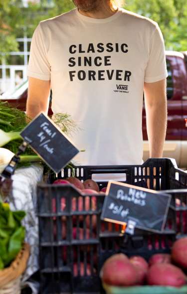 A vendor at the farmers' market standing behind their table of vegetables wearing a shirt that reads "Classic since forever"