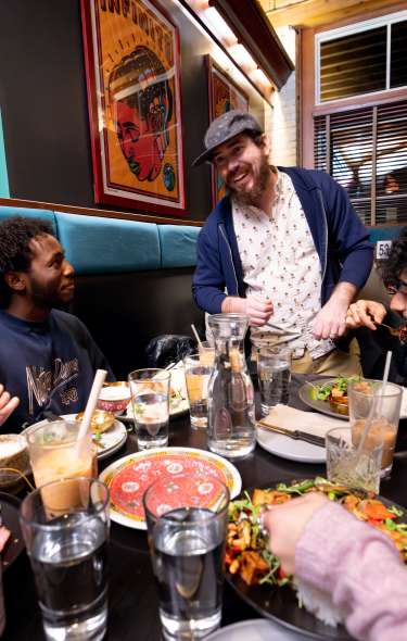 A group of diners gathered around a table in a cozy restaurant booth, sharing plates of food and drinks. The table is filled with colorful dishes, water glasses, and beverages, while warm lighting, wood accents, and framed artwork create an inviting atmosphere.