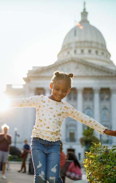 A young Black girl waves her arms like an airplane in front of the Capitol on a sunny day