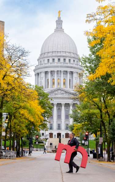 An image of a man walking across State Street in downtown Madison holding a large red "m" with the Capitol building in the background.
